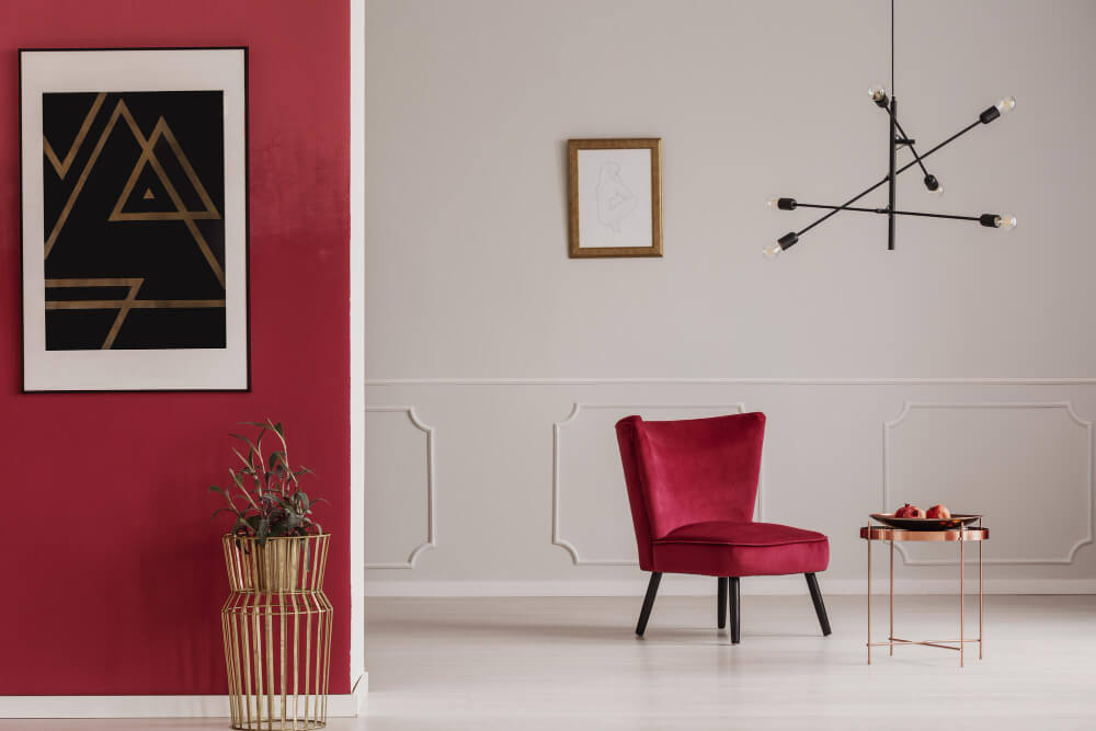 W nowoczesnym wnętrzu przedpokoju biała sztukateria ścienna kontrastuje z głęboką czerwienią ścian. Centralnym punktem jest elegancki czerwony fotel ustawiony na tle jasnej ściany z delikatnymi dekoracyjnymi panelami. Po lewej stronie widoczny jest złoty kwietnik z rośliną, a nad fotelem wisi czarna, nowoczesna lampa. Na podłodze znajduje się mały stolik kawowy w kolorze miedzi, który dopełnia stylową aranżację.